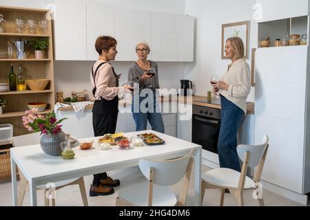 Reife Freunde unterhalten sich und trinken Wein mit Vorspeisen auf dem Tisch während des Treffens in der Küche Stockfoto