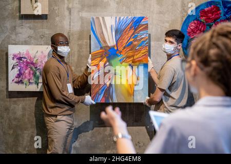 Zwei Arbeiter in der Galerie, die Handschuhe und Schutzmasken tragen, halten abstrakte Malerei und schauen auf eine Kollegin, während sie Kunstwerke an die Wand hängen Stockfoto