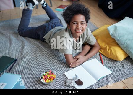 Glücklicher afrikanischer Junge, der Sie mit einem toothy Lächeln ansieht, während Sie auf dem Boden liegen, zeichnen, Süßigkeiten und Schokolade genießen und die Freizeit genießen Stockfoto