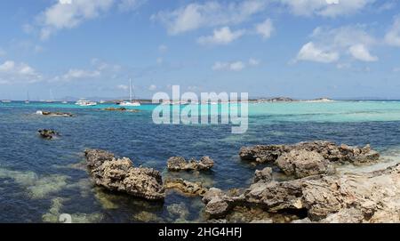 Panoramablick von einem felsigen Strand in Formentera, Spanien, im Hintergrund Segelboote vor dem blauen Himmel festgemacht. Stockfoto