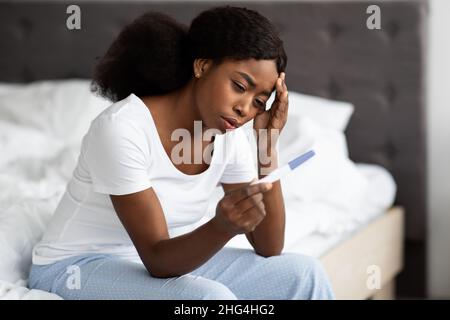 Rätselhafte schwarze Dame, die auf dem Bett sitzt und einen Schwangerschaftstest hält Stockfoto