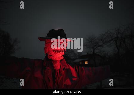 Schreckliche Vogelscheuche in dunklem Mantel und schmutzigen Hut beleuchtet mit rotem Licht steht in der Nähe des Hauses in der Nacht. Halloween-Konzept Stockfoto