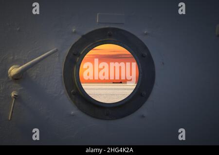 Einsames Fischerboot Trawler Boot auf dem Meerwasser. Blick vom runden Fenster aus der Hütte während des Sonnenuntergangs. Ruhiges klares Meer sonniges Wetter. Schöner Horizont der Seestücke Stockfoto