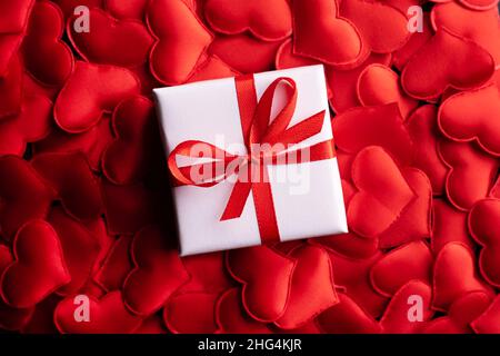 Kreatives Postkarten-Design am Valentinstag mit weißer Geschenkbox mit Schleife auf rotem Textilherzen-Hintergrund. Valentinstag und Liebeskonzept Stockfoto