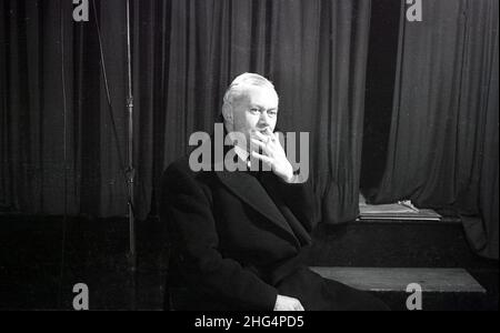 1950s, historisch, ein Mann, möglicherweise ein Schauspieler, sitzt in seinem Mantel an einer Bühne und Vorhänge mit einer Zigarette, England, Großbritannien. Stockfoto