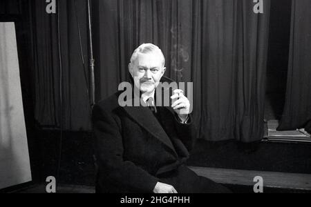 1950s, historisch, ein Mann, möglicherweise ein Schauspieler in Proben, sitzt in seinem Mantel an einer Bühne und Vorhänge in einer Pause, mit einer Zigarette, England, Großbritannien. Stockfoto