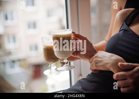 Die Hände des jungen Paares halten Glasküken mit heißem Latte-Kaffee. Liebhaber sitzen auf der Fensterbank am Fenster, trinken heiße Getränke in der Küche zu Hause i Stockfoto