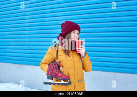 Die Skifahrerin in einer warmen, leuchtend gelben Jacke trinkt im Winter draußen Kaffee in der Nähe eines hellblauen Hintergrunds. Speicherplatz kopieren Stockfoto