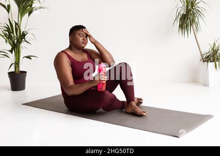 Erschöpfte übergewichtige schwarze Dame, die auf einer Fitnessmatte sitzt Stockfoto