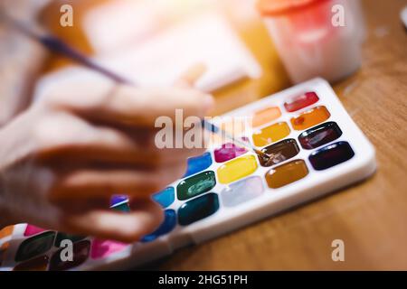 Hand mit Pinsel zum Zeichnen über mehrfarbige Palette von Aquarellfarben. Thema der Kreativität und Inspiration für Künstler. Hintergrund. Stockfoto