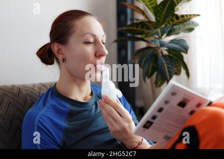 Eine kaukasische Frau liest ein Buch und hält einen Verneblerschlauch in der Hand, indem sie Medikamentendämpfe in ihre Lungen einatmet. Behandlung von Asthma oder kalter Grippe zu Hause. Inhalation bei Atemwegserkrankungen. Stockfoto