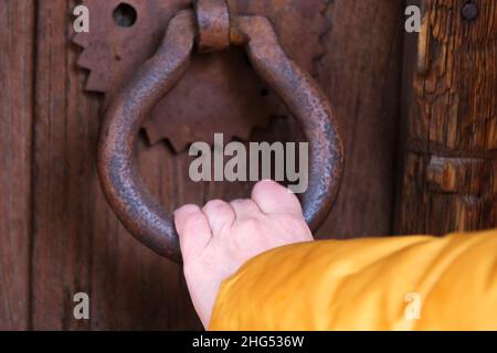 Weibliche Hand klopft mit uraltem eisernem Türknauf an die Tür, Frau öffnet die Holztür mit bronzeverrostem Griff. Stockfoto