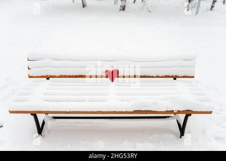 Auf einer Holzbank liegt ein rotes Rattan-Herz auf dem weißen Schnee vor der Kulisse eines verschneiten Waldes. Urlaubskarte mit Liebe. Einsames, winterliches Herz. Kopieren Stockfoto