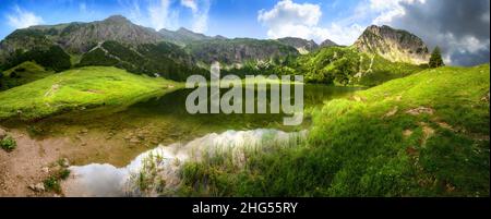 Bergseen-Panorama mit grünen Wiesen, blauem Himmel, dramatischen Wolken und Reflexionen auf das klare Wasser der Alpen Stockfoto