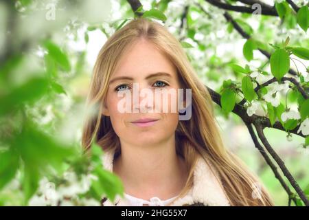 Porträt einer schönen jungen Frau in der Natur, mit Frühlingsblüten und zartem grünem Laub auf Ästen, die ihr Gesicht umgaben Stockfoto