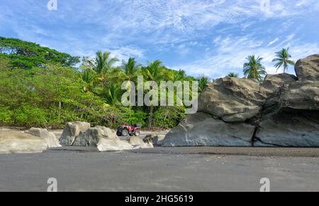 Low-Angle-Ansicht eines roten Geländefahrrads, das auf einem felsigen Strand von Costa Rica vor Palmen und blauem Himmel vor Hintergrund steht. Stockfoto