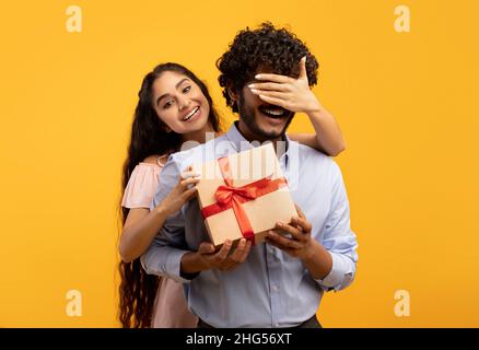 Hübsche indische Frau bedeckt die Augen ihres Freundes, hält eine Geschenkbox und begrüßt ihn mit Geburtstag oder Jahrestag Stockfoto