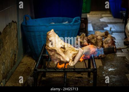 Bangkok, Thailand. 18th Januar 2022. Der Schweineköpfen wird auf einem Grill geröstet, bevor er auf einem Markt verkauft wird.Arbeiter am Großmarkt Bangkok Noi bereiten am 18. Januar 2022 frisch geschlachtetes Schweinefleisch zum Verkauf vor. Aufgrund von Lieferengpässen, Marktspekulationen und einem möglichen Ausbruch der Afrikanischen Schweinepest steigen die Schweinefleischkosten in Thailand in die Höhe, was den Preis für Grundnahrungsmittel in die Höhe schiessen lässt. Die schnelle Inflation hat zu einem plötzlichen landesweiten Anstieg von Lebensmitteln von der Straße bis zum Supermarkt geführt. Kredit: SOPA Images Limited/Alamy Live Nachrichten Stockfoto