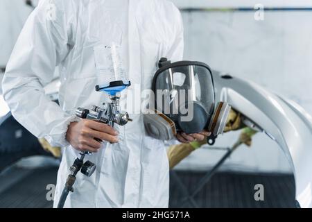 Сlose-up-Ansicht des Automalers mit Farbpistole und Schutzmaske in den Händen. Reparaturkonzept für das Auto. Mechaniker in der Lackierkabine des Fahrzeugs Stockfoto