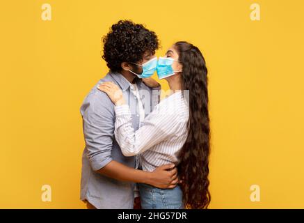 Liebe während einer Pandemie. Das junge indische Paar küsst und umarmt sich und trägt medizinische Gesichtsmasken auf gelbem Hintergrund Stockfoto