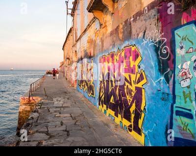 Lissabon, Portugal - 18. Oktober 2021: Graffiti und Wandmalereien in der Abenddämmerung auf verlassenen Gebäuden im Lissabonner Almada-Viertel am Wasser o Stockfoto