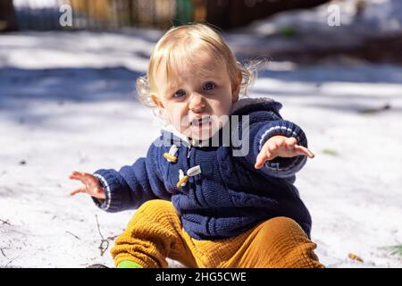 Vorderansicht eines 3-jährigen Jungen mit besorgtem Gesichtsausdruck, weiten Augen und offenem Mund, der die Hand zur Kamera reicht, die auf Schnee mit verschwommenem Hintergrund sitzt. Stockfoto