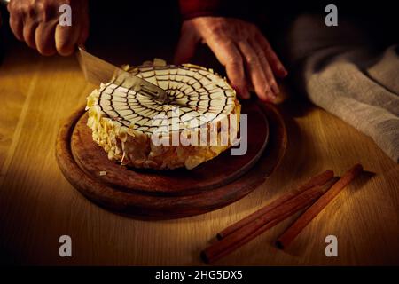 Eine Frau schneidet einen Esterhazy Mandel-Schokoladenkuchen auf einem Brett mit einem Holztisch im Hintergrund. Stockfoto