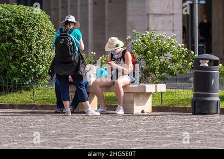 Pisa, Italien - 10. August 2021: Menschen mit Gesichtsmasken sitzen auf einer Bank im Park in der Nähe des Bahnhofs und nutzen ein Smartphone Stockfoto