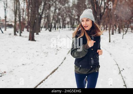 Junge Sportlerin, die im verschneiten Winterpark unter fallendem Schnee läuft. Aktiver, gesunder Lebensstil. Workout im Freien Stockfoto