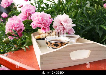 Tee im Landhausstil im Sommergarten im Dorf. Zwei Tassen heißen schwarzen Tee auf einem Holztablett und blühende rosa Pfingstrosen im Sonnenlicht. Stockfoto