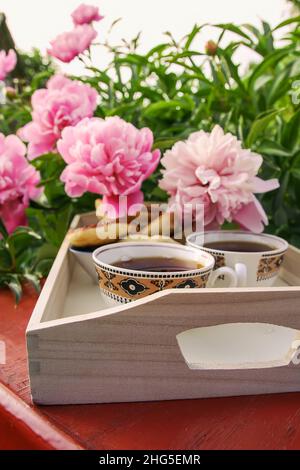 Tee im Landhausstil im Sommergarten im Dorf. Zwei Tassen heißen schwarzen Tee auf einem Holztablett und blühende rosa Pfingstrosen im Sonnenlicht. Stockfoto