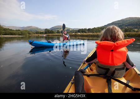 Das junge, siebenjährige Mädchen trägt eine Rettungsweste vor einem Kajak für zwei Personen, während der Mann auf windermere im Sommer-Seengebiet in cumbria, en, vorbeipaddelt Stockfoto