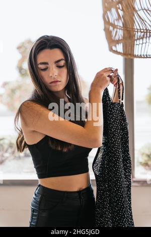 Junge Brünette in legerer Kleidung mit trendigen Kleid, während sie sich am sonnigen Tag zu Hause gegen das Fenster kleiden Stockfoto