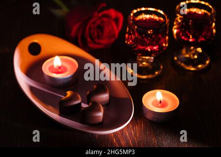 Herzförmige Schokoladenpralinen mit Kirschlikör in zwei Gläsern mit roter Rose und Kerzen in romantischer Umgebung. Valentinstag-Konzept. Romantisch. Stockfoto