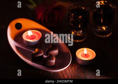 Herzförmige Schokoladenpralinen mit Kirschlikör in zwei Gläsern mit roter Rose und Kerzen in romantischer Umgebung. Valentinstag-Konzept. Romantisch. Stockfoto