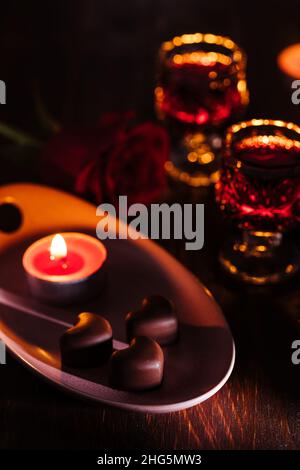 Herzförmige Schokoladenpralinen mit Kirschlikör in zwei Gläsern mit roter Rose und Kerzen in romantischer Umgebung. Valentinstag-Konzept. Romantisch. Stockfoto