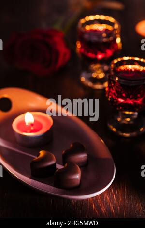 Herzförmige Schokoladenpralinen mit Kirschlikör in zwei Gläsern mit roter Rose und Kerzen in romantischer Umgebung. Valentinstag-Konzept. Romantisch. Stockfoto