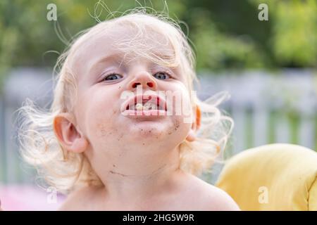 Nahaufnahme ein lustiges Porträt eines dreijährigen Jungen mit humorvoller Persönlichkeit, die mit Essen aus dem Mund ragt und schmutzigem Gesicht posiert. Mit verschwommenem Hintergrund. Stockfoto