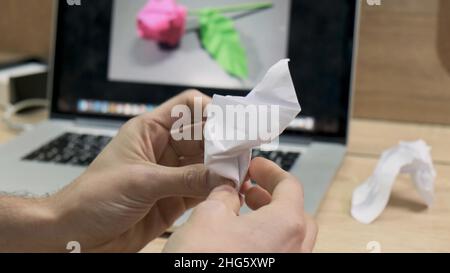 Männliche Hände machen Origami-Figur aus weißem Papier. Der Mensch macht aus Papier keine Blume wie auf einem Bild auf einem Computerbildschirm, Stockfoto