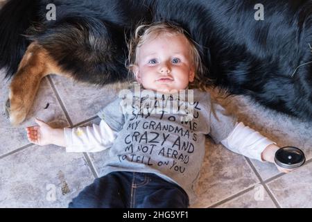 High-Angle-Aufnahme eines dreijährigen Jungen, voller Unfug, der auf einem gefliesten Küchenboden lag und Kopf auf dem Bauch eines großen Familienhundes mit Kopieplatz lag. Stockfoto