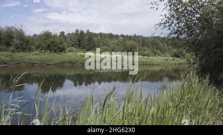 Wunderschöne Sommerlandschaft mit dem Fluss und seinen grünen Ufern und einem Mann, der in einem aufblasbaren Boot schwimmt. Fischer Reiten aufblasbare Gummiboot auf der Stockfoto