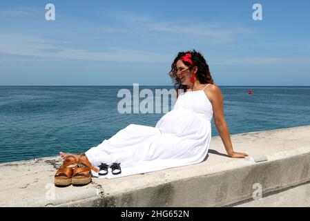 Eine Schwangerin sitzt entspannt am Meer und genießt die frische Luft, die Sonne und den Meerblick. Ihre Schuhe neben ihren erwarteten Babys Stockfoto