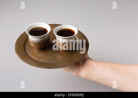 Eine Männerhand hält ein Kupfertablett mit zwei orientalischen Kaffeetassen. Frisch gebrühter türkischer Kaffee in Tassen. Das Konzept des Minimalismus. Stockfoto