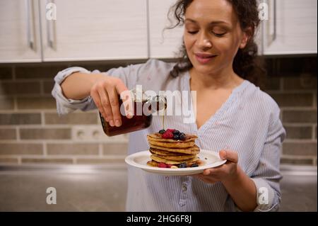 Konzentrieren Sie sich auf die Hände der verschwommenen schönen Köchin, der charmanten hübschen Frau, die Ahornsirup auf einen Stapel hausgemachter Pfannkuchen mit Beeren gießt Stockfoto