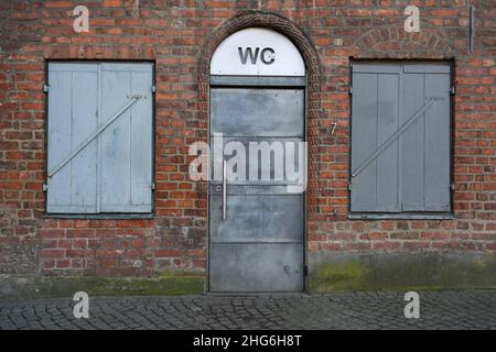 Öffentliche Toilette mit einer grauen Metalltür und einem Schild WC in einem historischen roten Backsteingebäude in der Altstadt von Lübeck, Deutschland Stockfoto