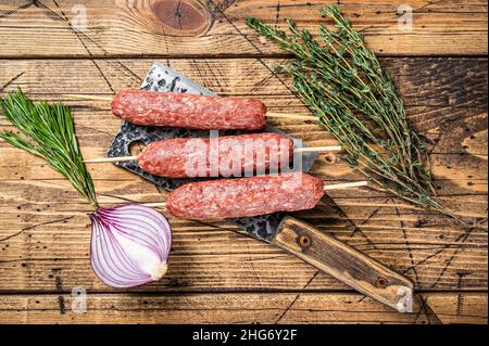Frische rohe Kofta oder lula Kebabs Spieße auf einem Metzgerspalter mit Kräutern. Holzhintergrund. Draufsicht Stockfoto