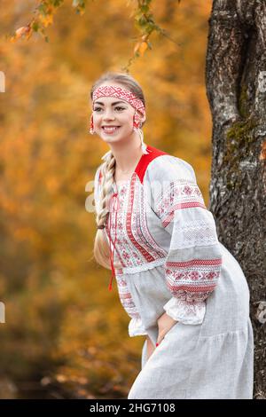 Junge attraktive Frau lächelt in volkstümlicher traditioneller slawischer Kleidung in der Nähe des Baumes auf die Natur im Herbstwald. Portrait von schönen Mädchen in ethnischen Kleidung Stockfoto