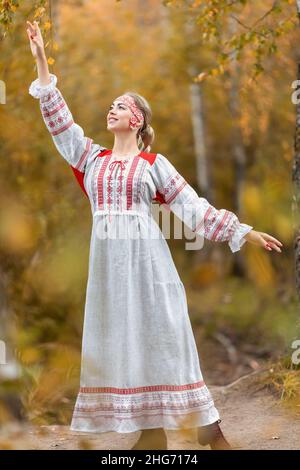 Portrait der jungen schönen lächelnden Frau in volkstümlichen traditionellen slawischen Kleidern grüßt Naturgeister von Hand im Herbstwald. Stockfoto