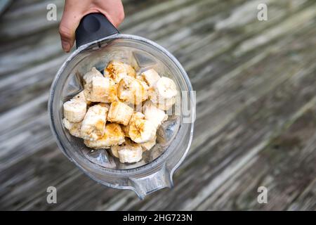 Flache Nahaufnahme der Hand mit gefrorenen geschälten gelben Bananen in Kunststoff-Mixerbehälter für Obst hausgemachten Smoothie oder vegane nicht-Milchprodukte schöne Eiskre Stockfoto