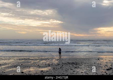 Am Sonntag, den 16. Januar 2022, läuft eine junge Frau am Strand in Oceanside, Oregon, bei Sonnenuntergang vor der Welle. Die Tsunami-Bedrohung im Pazifik wird durch eine... Stockfoto